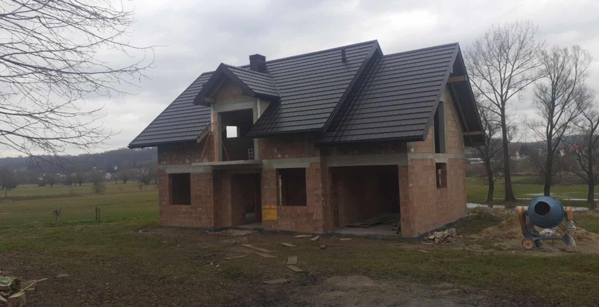 Niedokończony dom jednorodzinny z ciemną dachówką, widoczna murowana konstrukcja ścian i nieotynkowane ściany szczytowe, na pierwszym planie betoniarka.