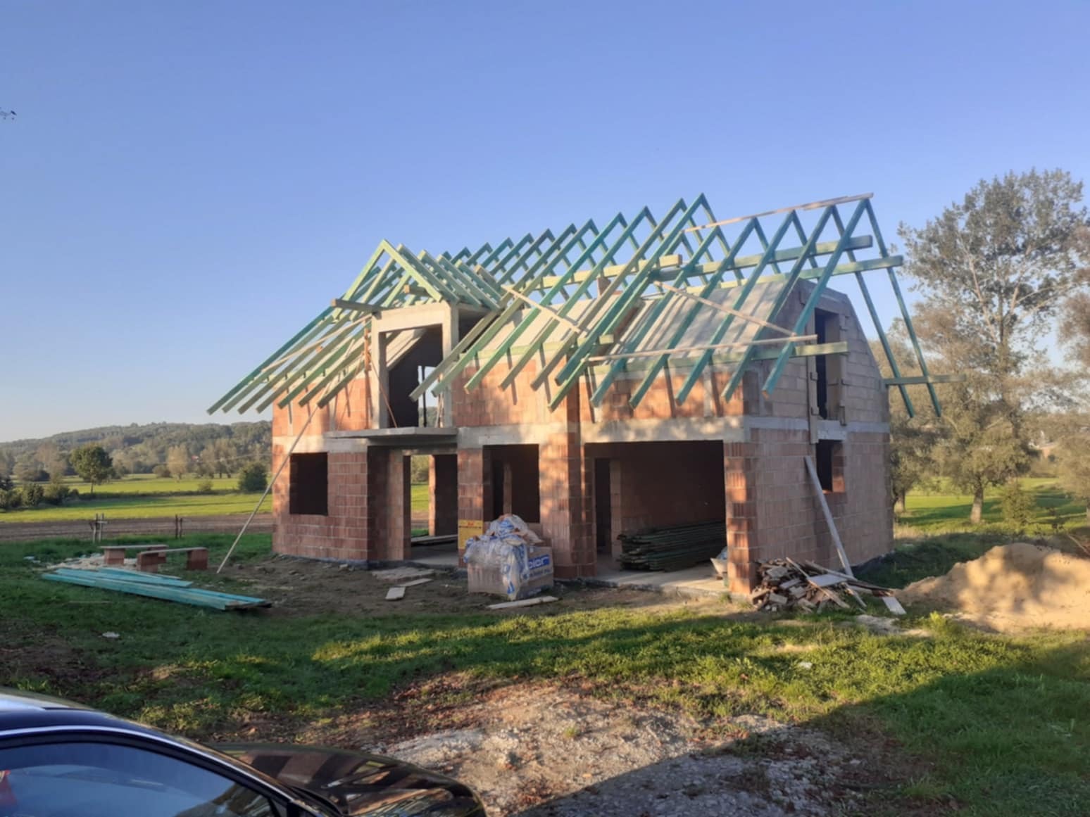 Surowy budynek jednorodzinny z czerwonej cegły w trakcie budowy, widoczne ściany i drewniana konstrukcja dachu na tle zielonego krajobrazu.