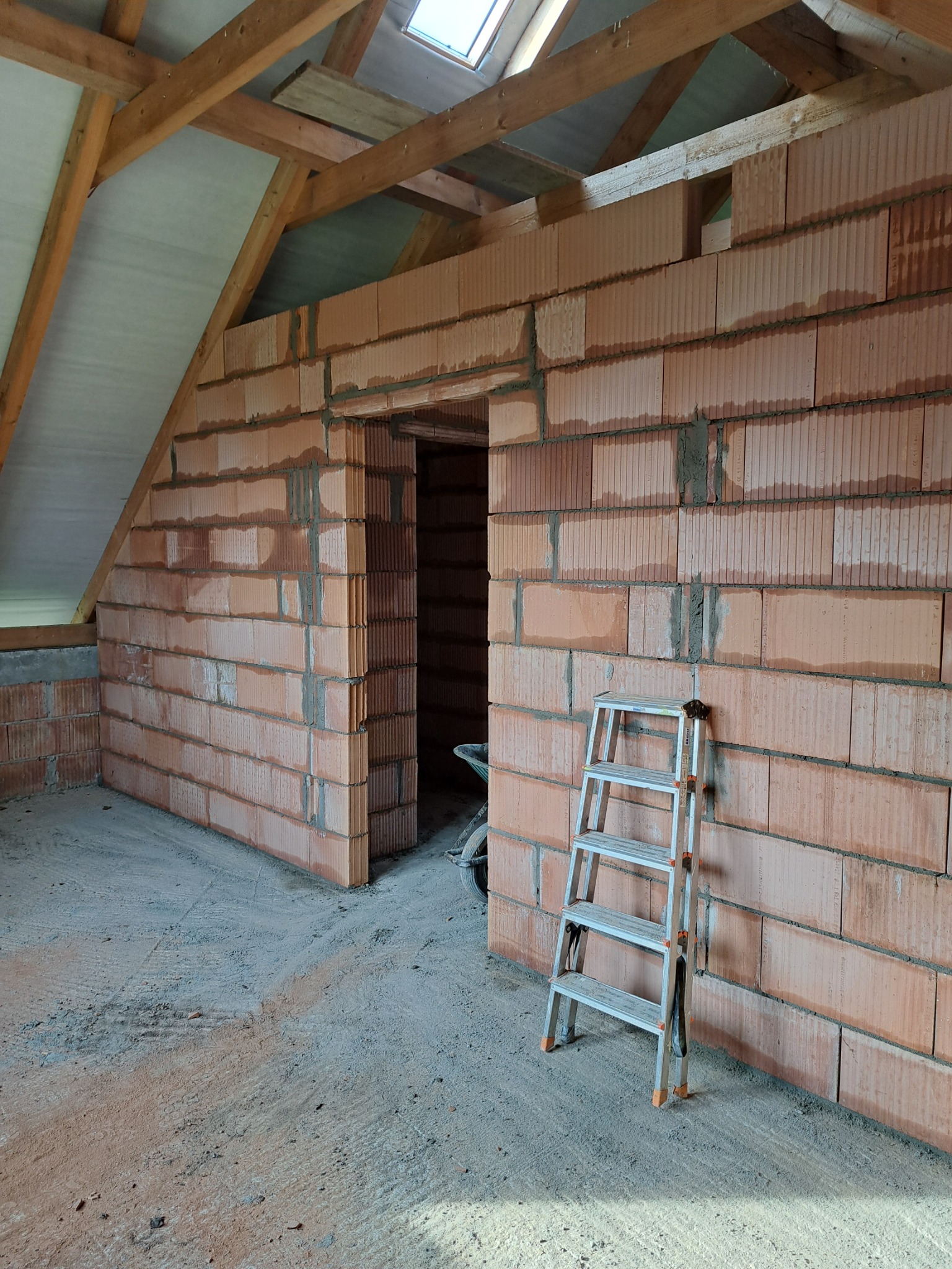 Wnętrze domu w Niedźwiadzie w trakcie budowy, widoczne ściany z czerwonej cegły, drabina aluminiowa oparta o ścianę oraz drewniana konstrukcja dachu z oknem dachowym.