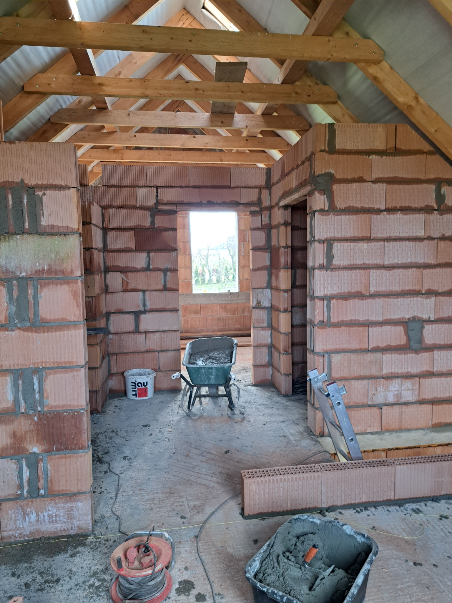 Wnętrze budynku w trakcie budowy, widoczne ściany z czerwonej cegły, drewniana konstrukcja dachu, taczka z zaprawą, wiadro z klejem i zwinięty przedłużacz.