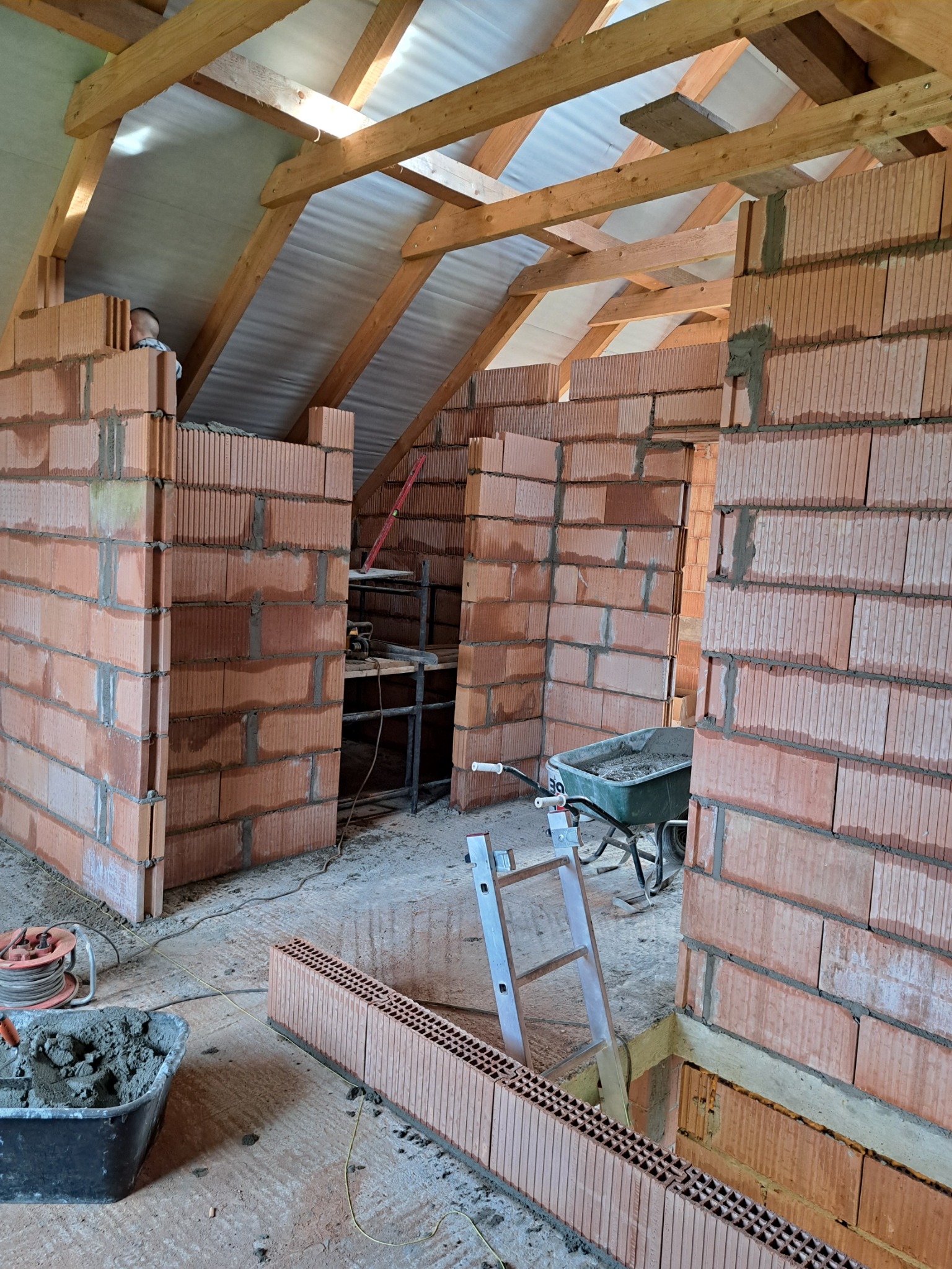 Wnętrze budynku w trakcie wznoszenia ścian z czerwonej cegły ceramicznej, widoczne drewniane elementy konstrukcji dachu, taczka z zaprawą, drabina, oraz narzędzia murarskie.
