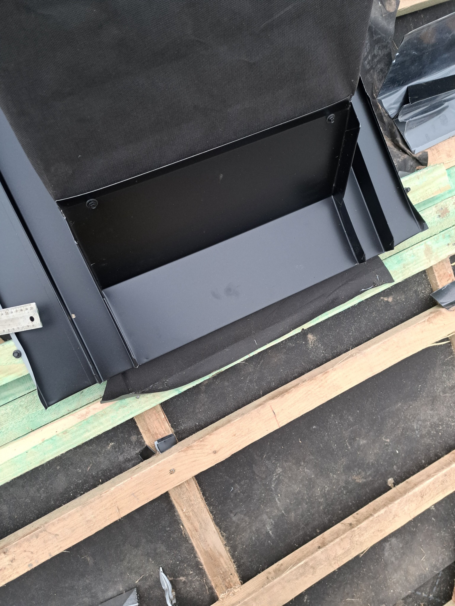 Montaż czarnej obróbki blacharskiej komina na dachu z widoczną łatą i kontrłatą, z użyciem membrany dachowej i narzędzi dekarskich.