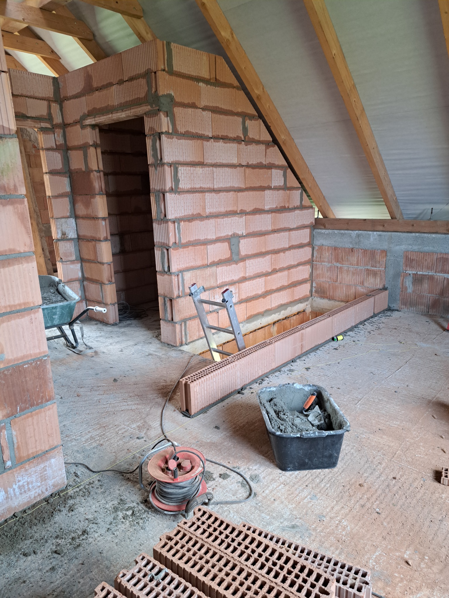 Wnętrze poddasza w trakcie budowy ścian z czerwonej cegły ceramicznej, widoczne narzędzia i materiały budowlane, surowe wykończenie, drewniana konstrukcja dachu w tle.