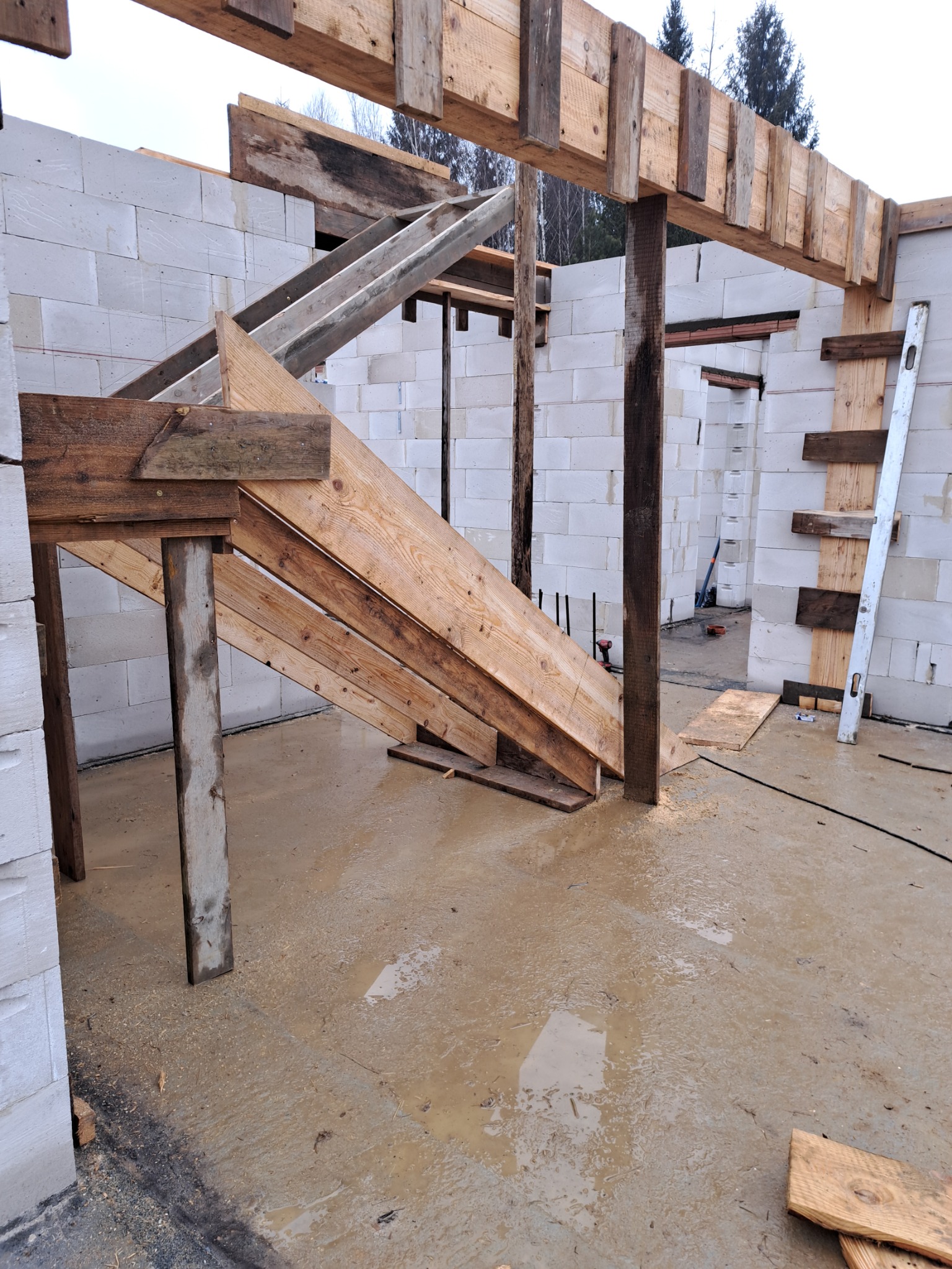 Wznoszenie ścian z bloczków betonu komórkowego, widoczne drewniane szalunki i podpory, mokra podłoga wewnątrz budynku w trakcie budowy, poziomica oparta o ścianę.