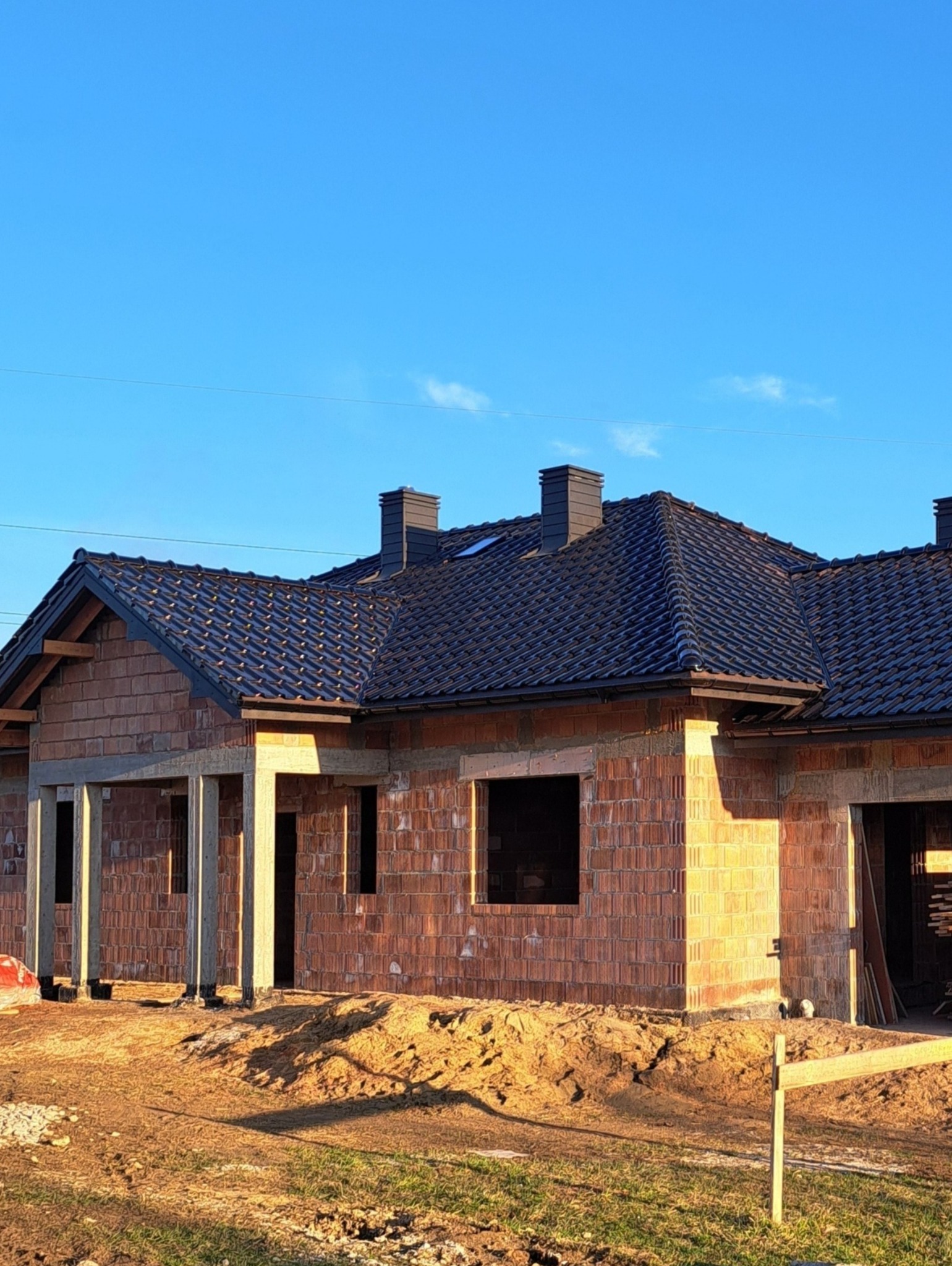 Surowy stan domu z cegły ceramicznej z ciemną dachówką i kominami w słoneczny dzień. Widoczne otwory okienne i drzwiowe, teren wokół w stanie budowy.