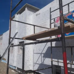 Budynek w trakcie ocieplania styropianem, widoczny fragment rusztowania i materiały izolacyjne z naklejonymi etykietami producenta, surowy teren wokół