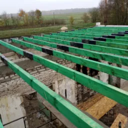 Wiązary dachowe pomalowane na zielono, ułożone na murach starego budynku w trakcie renowacji, z widocznymi elementami izolacji i krajobrazem w tle.