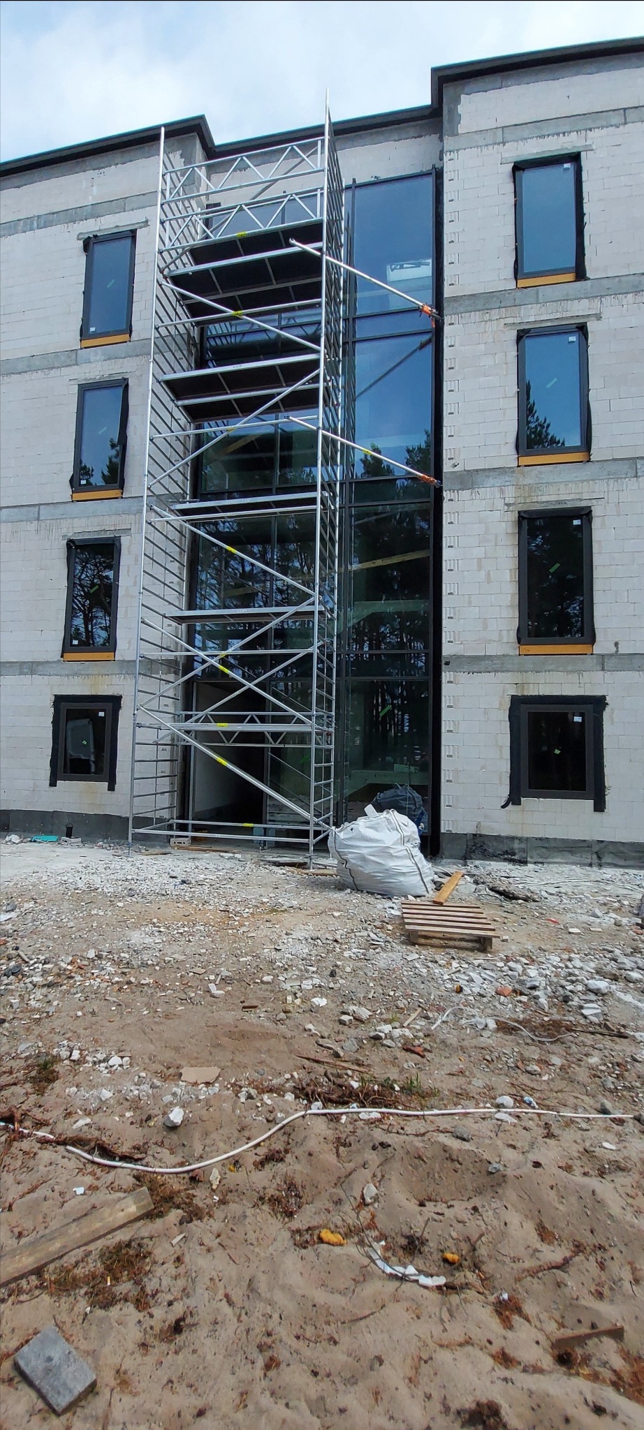 Wielopiętrowy budynek z białego bloczka w trakcie montażu okien, z widocznym rusztowaniem i workiem z odpadami budowlanymi na pierwszym planie.