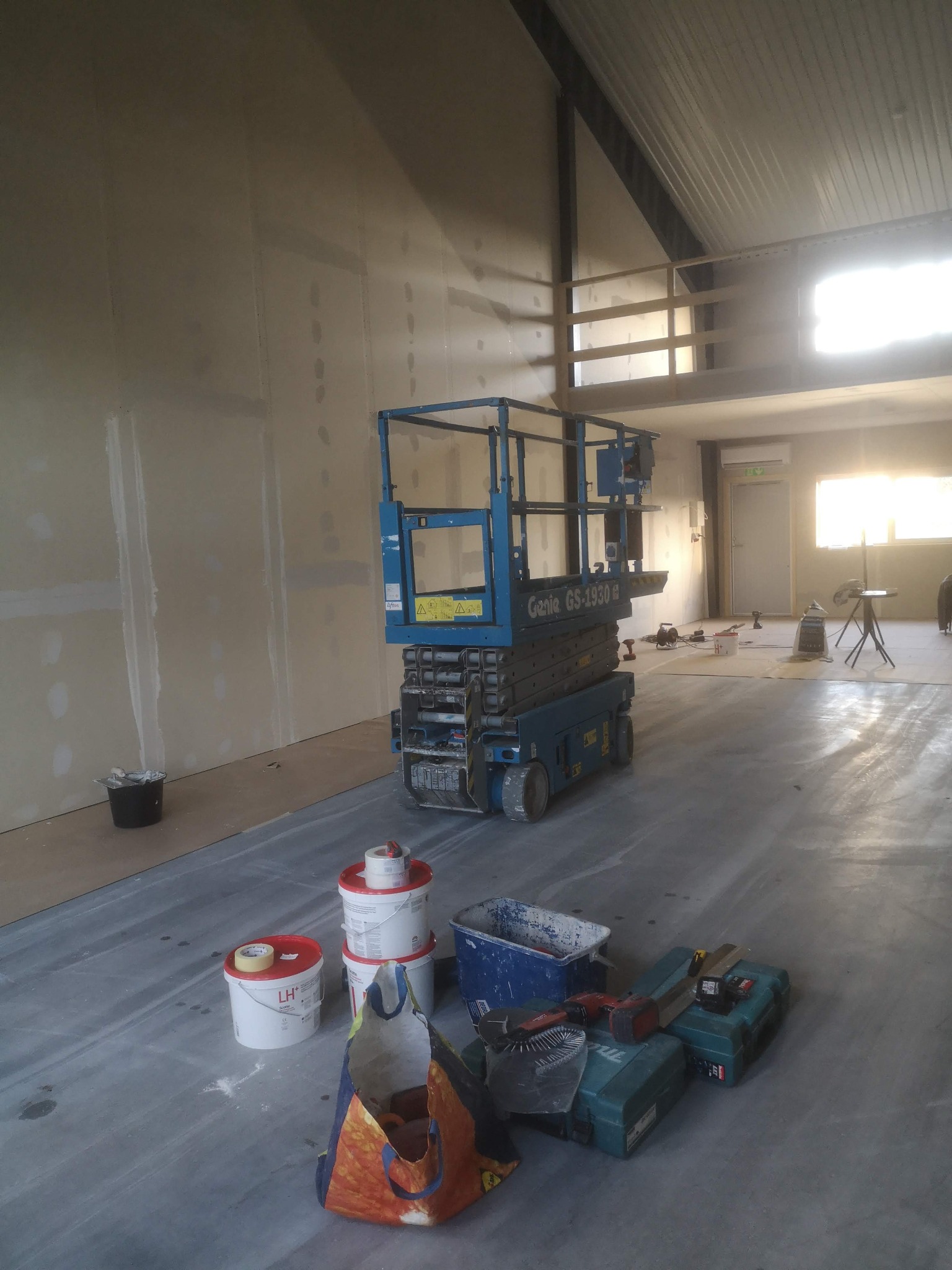 Wnętrze w trakcie prac wykończeniowych z podnośnikiem Genie GS-1930, widoczne ściany z płyt kartonowo-gipsowych przed szpachlowaniem, narzędzia i puste wiadra po farbach na betonowej podłodze.