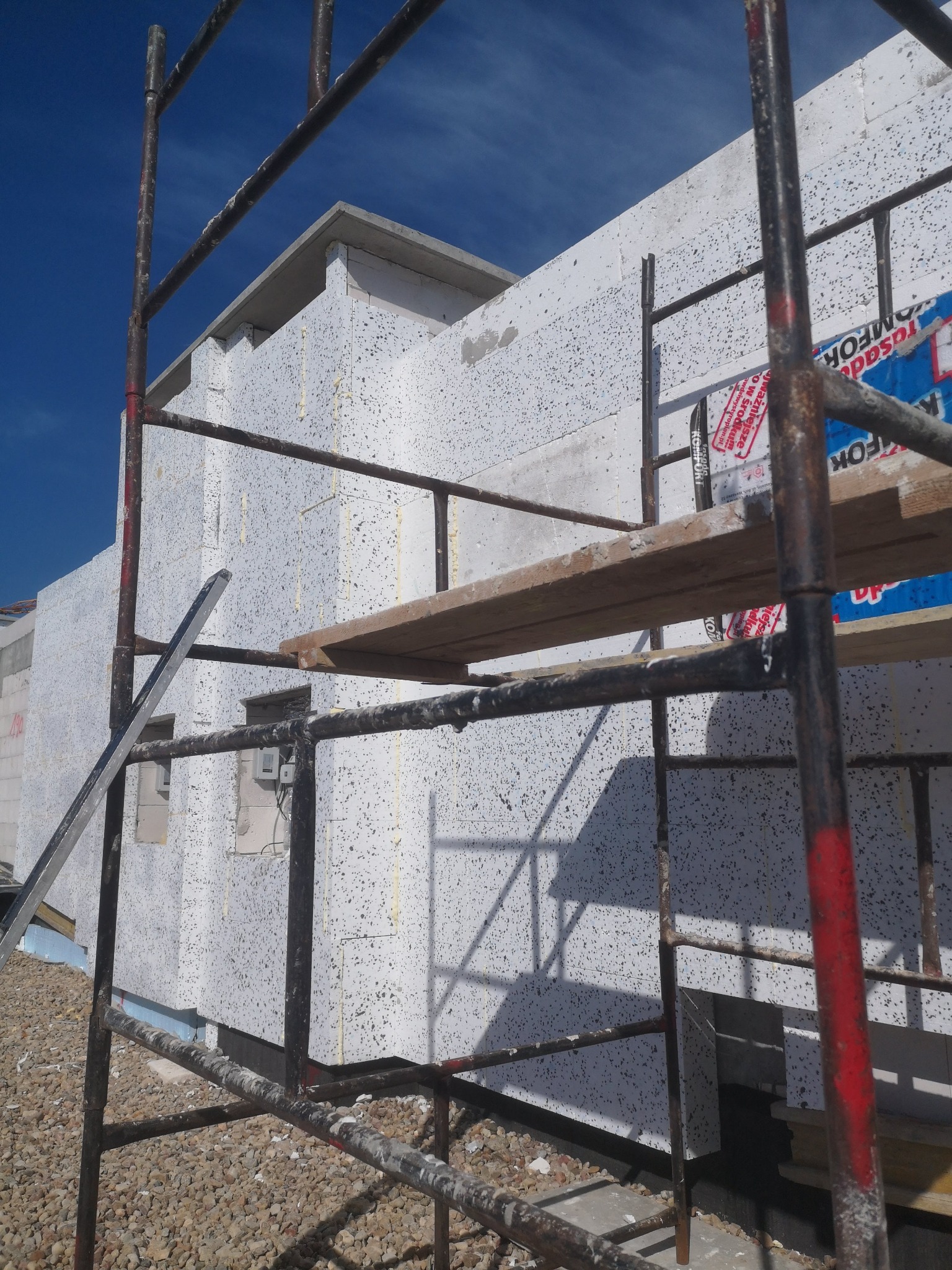 Budynek w trakcie ocieplania styropianem, widoczny fragment rusztowania i materiały izolacyjne z naklejonymi etykietami producenta, surowy teren wokół