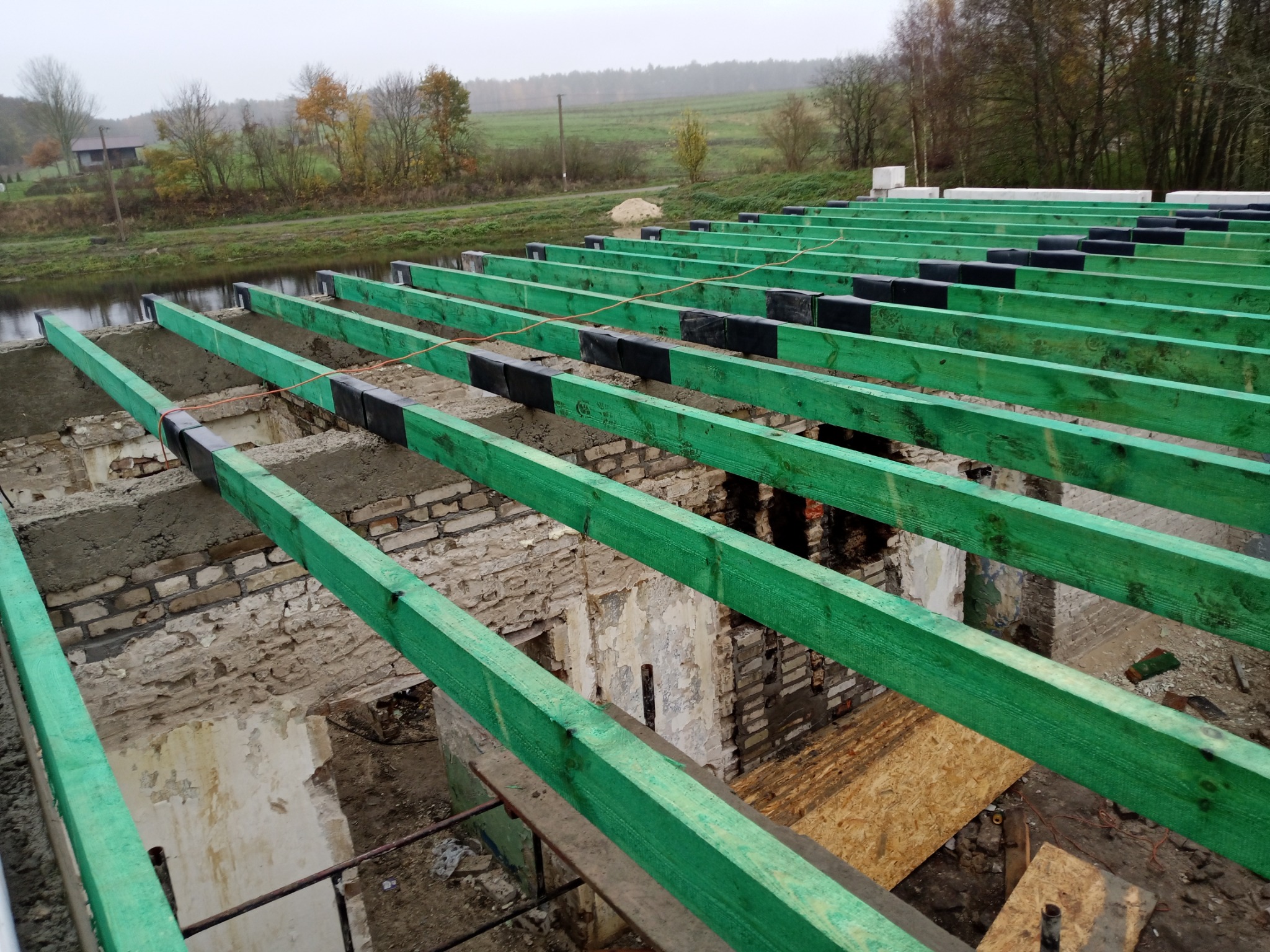 Wiązary dachowe pomalowane na zielono, ułożone na murach starego budynku w trakcie renowacji, z widocznymi elementami izolacji i krajobrazem w tle.