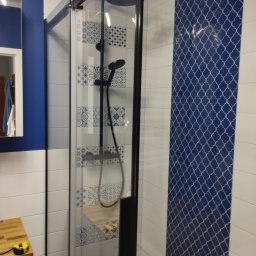 Remont - Kabina prysznicowa z czarnymi profilami, wykończona płytkami w odcieniach bieli i granatu z dekoracyjnymi wzorami, widoczny blat z żółtą miarką.