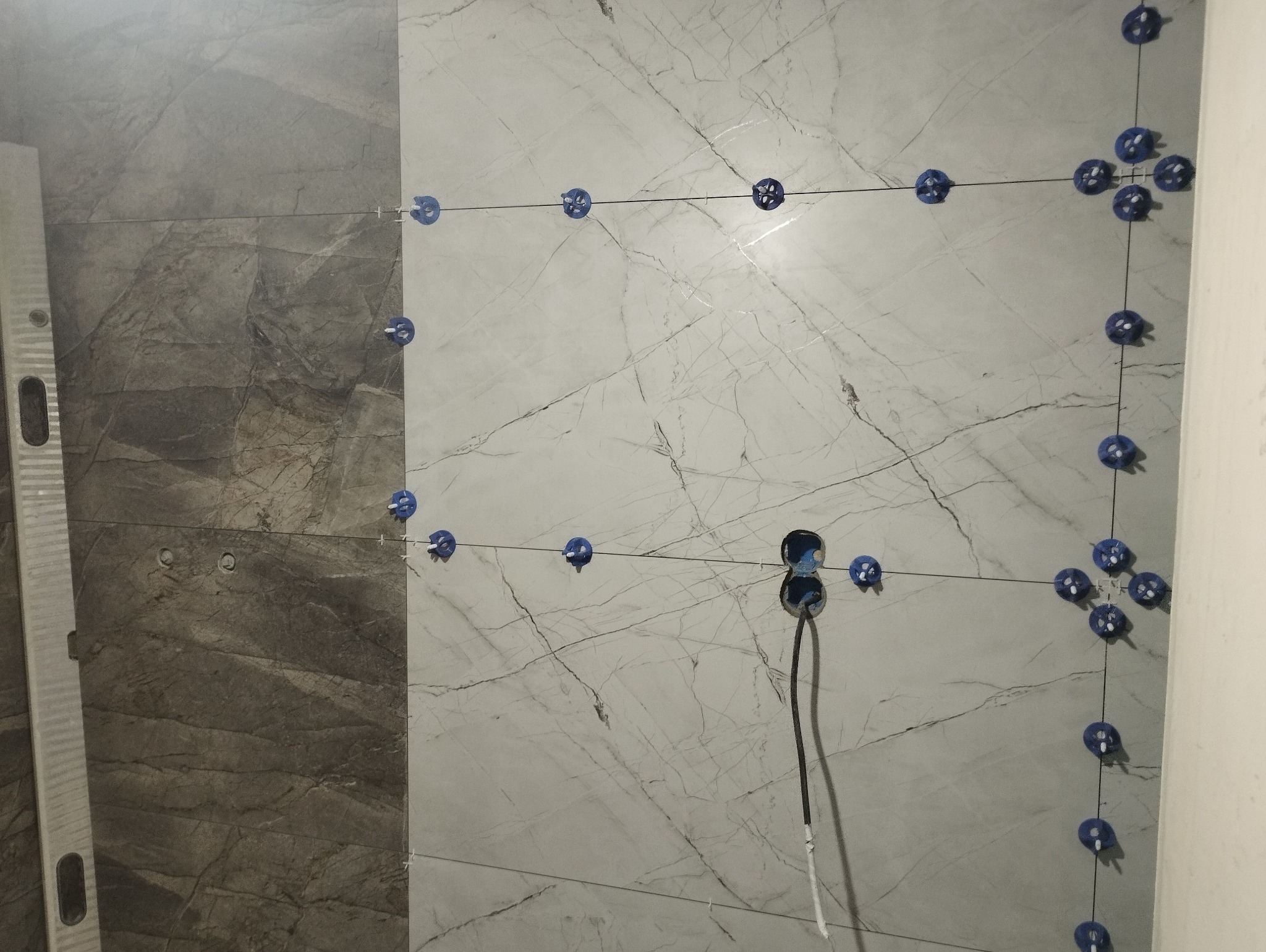 Montaż płytek imitujących marmur na ścianie, z użyciem systemu poziomowania z niebieskimi klipsami i sznurkami, widoczny poziomica przy ścianie i otwór na gniazdko z wystającym przewodem.