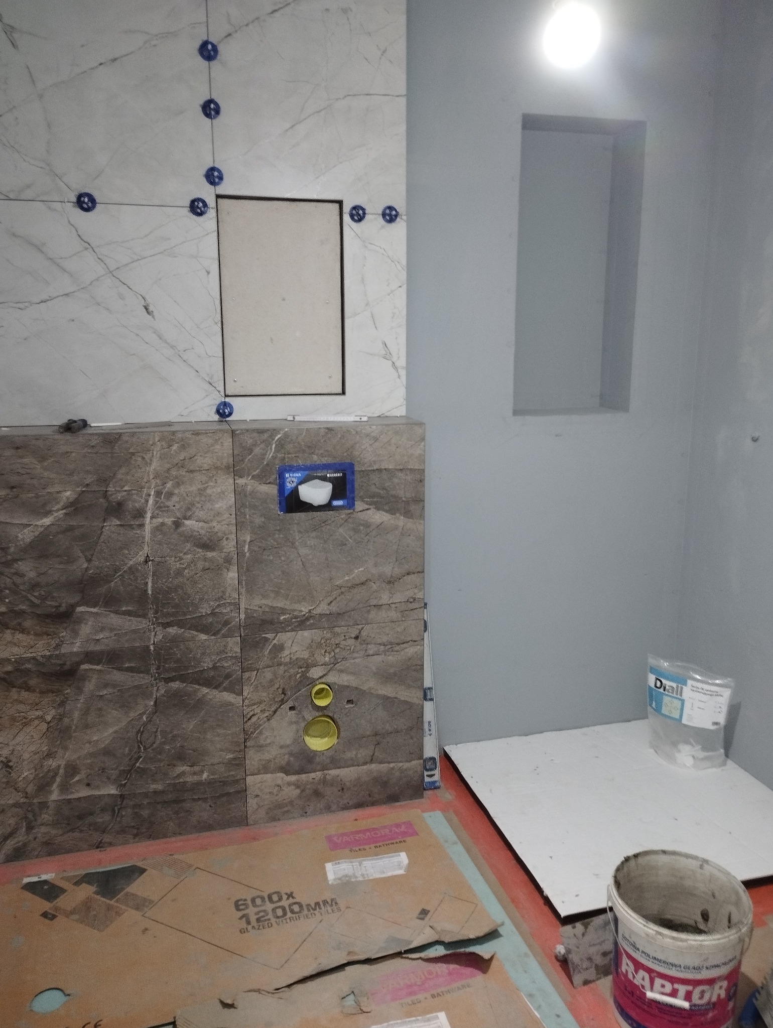Fragment łazienki w trakcie prac: białe i szare płytki imitujące marmur na ścianach, niebieskie krzyżyki dystansowe, otwory na instalacje, paczka kleju Diall oraz wiadro po kleju Raptor na podłodze...