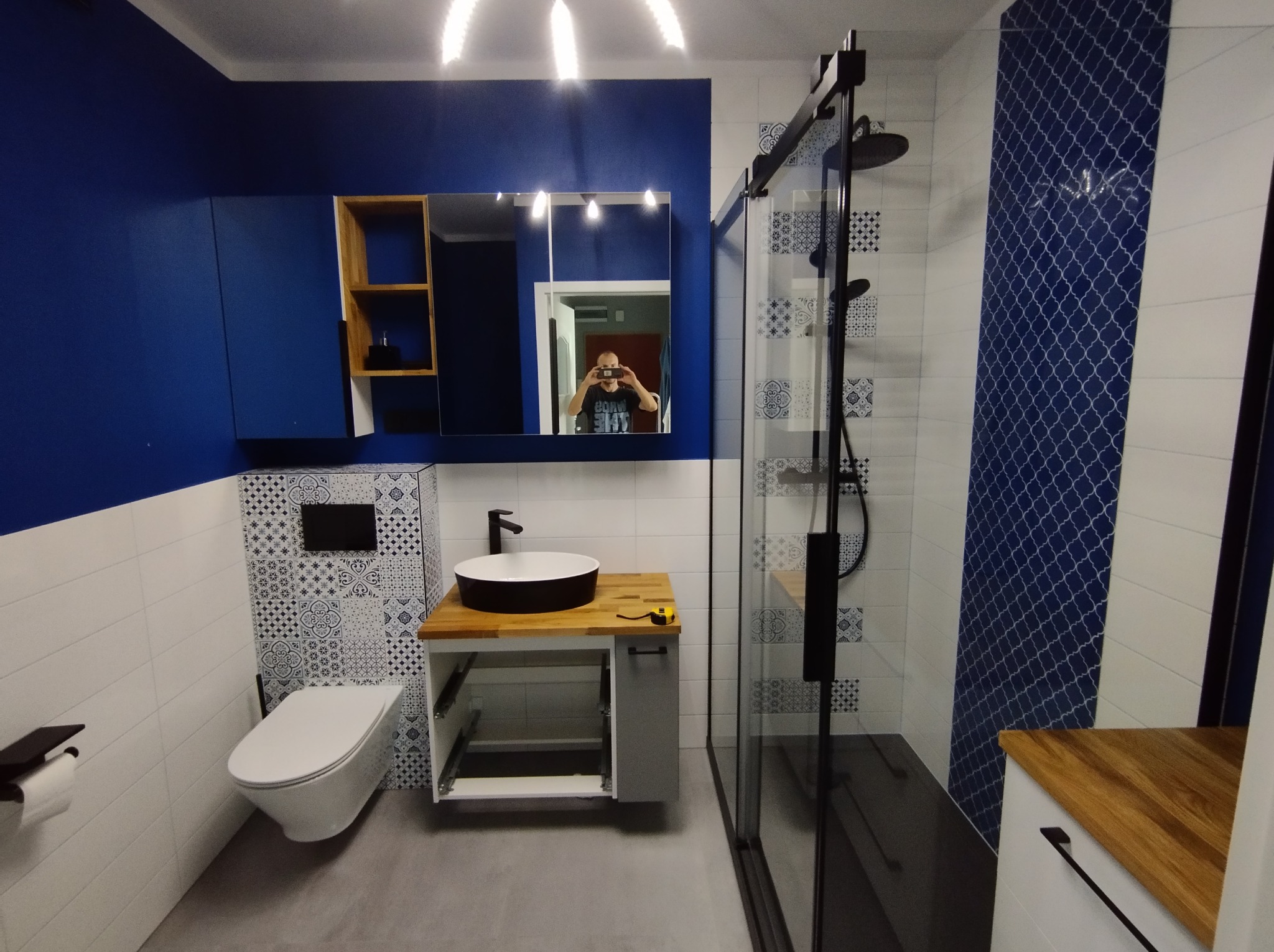 Nowoczesna łazienka w bieli i granacie z kabiną prysznicową typu walk-in, umywalką nablatową na drewnianym blacie i podwieszaną toaletą; osoba robiąca zdjęcie widoczna w lustrze.