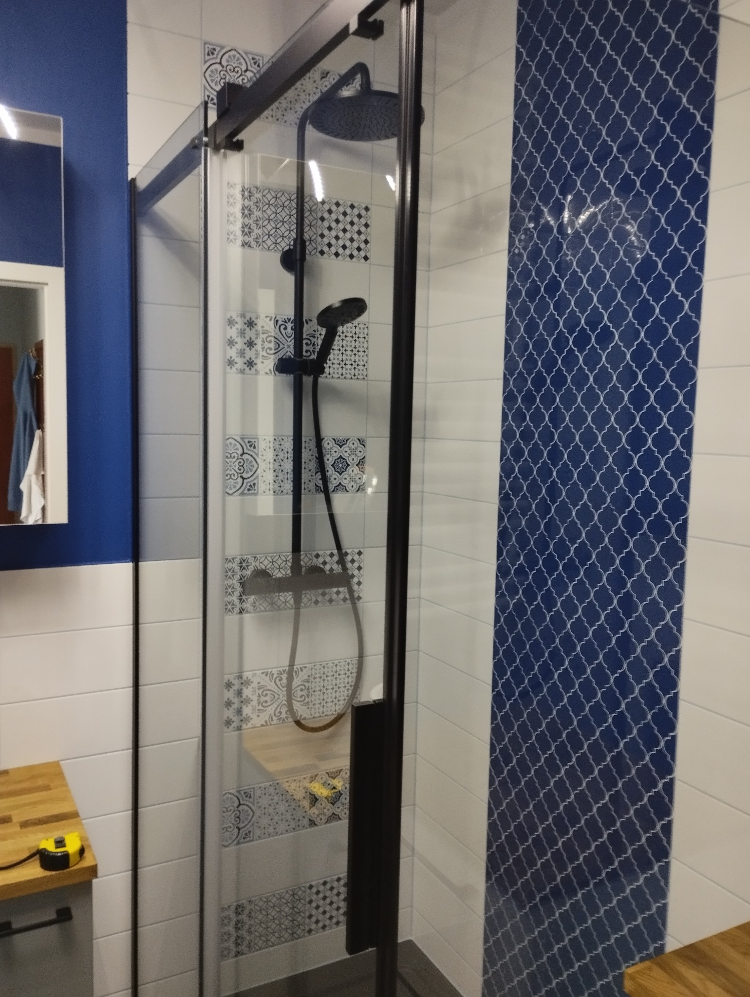 Kabina prysznicowa z czarnymi profilami, wykończona płytkami w odcieniach bieli i granatu z dekoracyjnymi wzorami, widoczny blat z żółtą miarką.