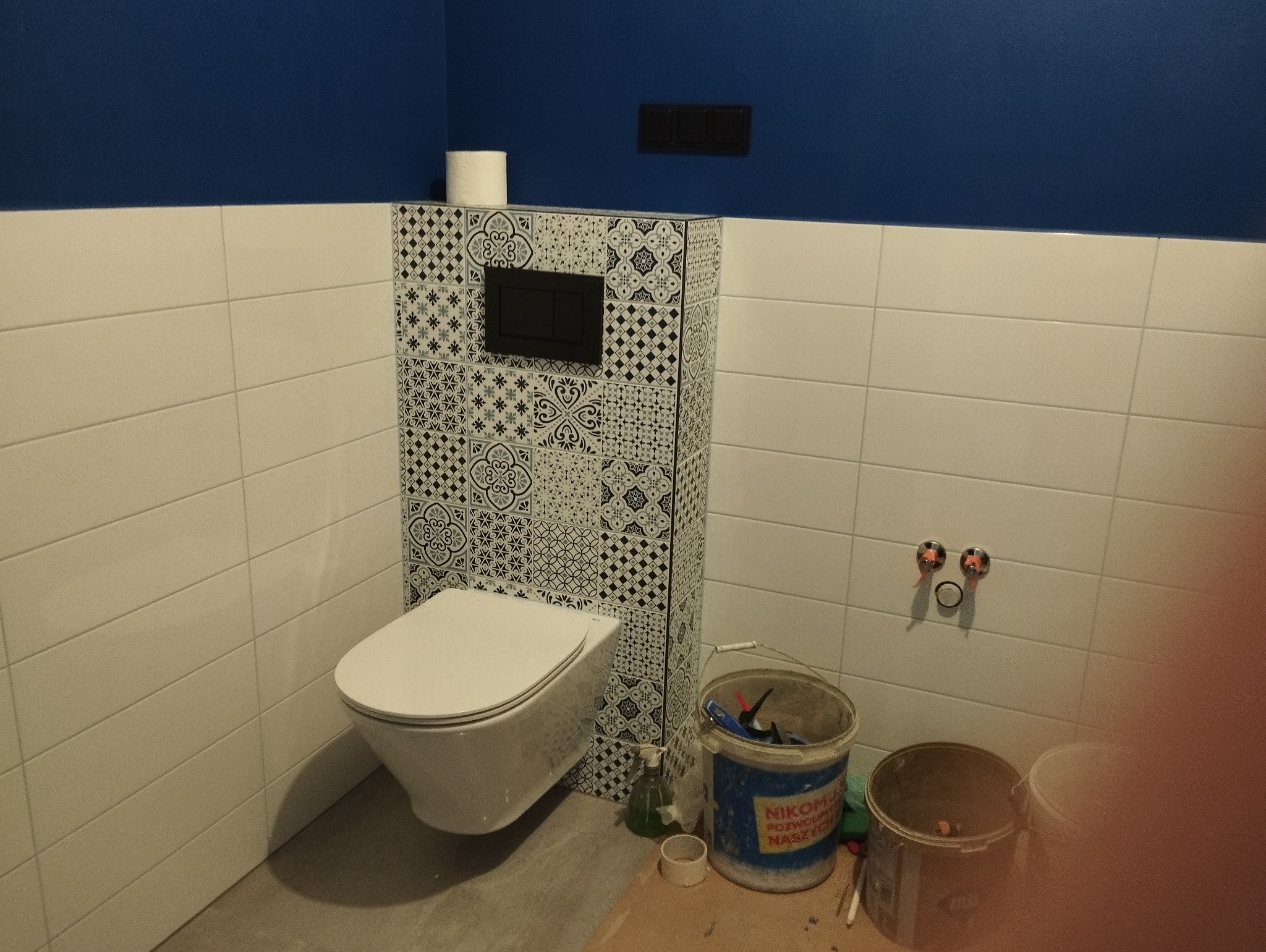 Podwieszana toaleta z ozdobną zabudową w czarno-białe wzory, zamontowana w łazience w Szczecinie. Widoczne białe płytki na ścianach i niebieska góra ściany. U dołu wiadra i narzędzia.