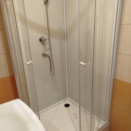 DC INSTAL Dominik Cech - Kabina prysznicowa z brodzikiem i armaturą, widok z boku, częściowo widoczna umywalka. Beżowe płytki na ścianach. Zamontowana słuchawka prysznicowa.