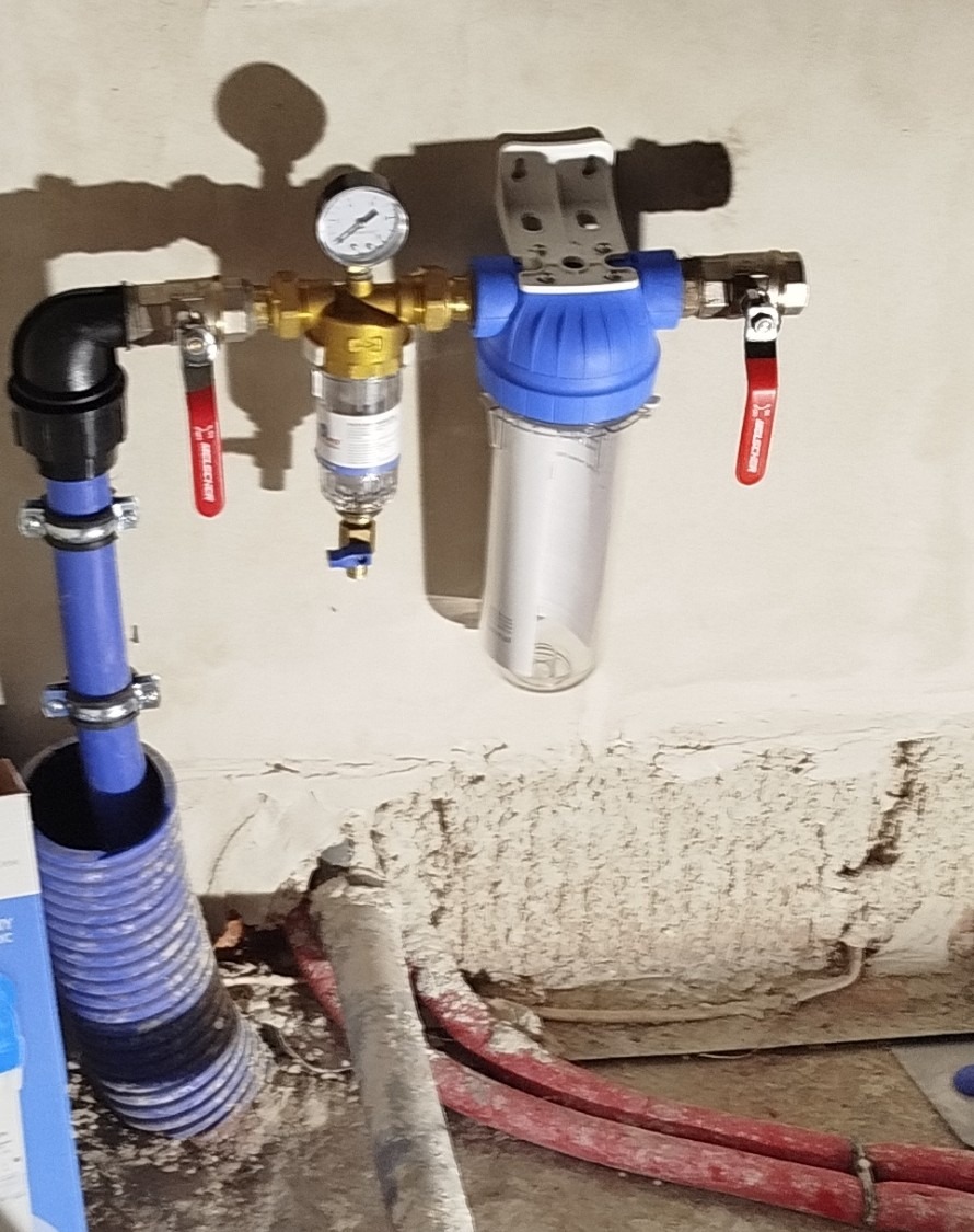 Zamontowany filtr wody z manometrem i zaworami odcinającymi na tle surowej ściany. Widoczne rury instalacyjne i fragment podłogi w trakcie prac wykończeniowych.