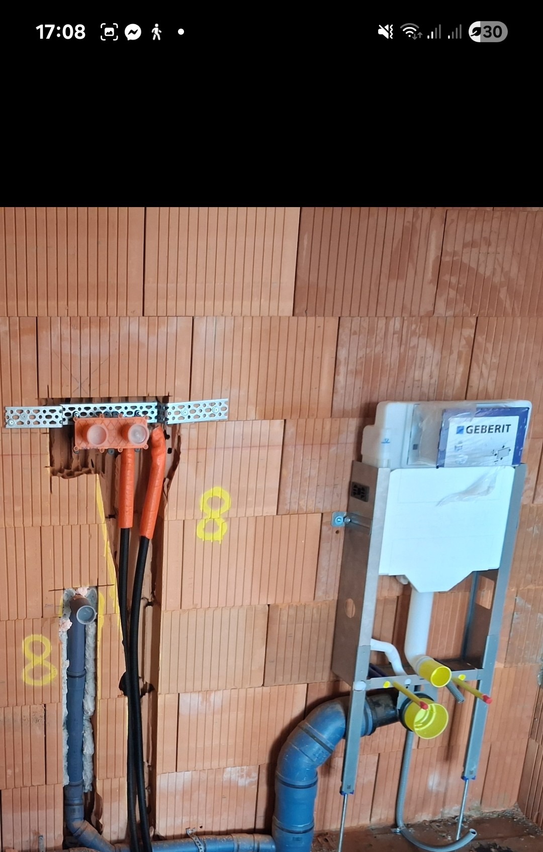 Ściana z cegieł z zamontowaną instalacją podtynkową WC Geberit, widoczne rury kanalizacyjne i doprowadzenie wody. Etap prac instalacyjnych w łazience.