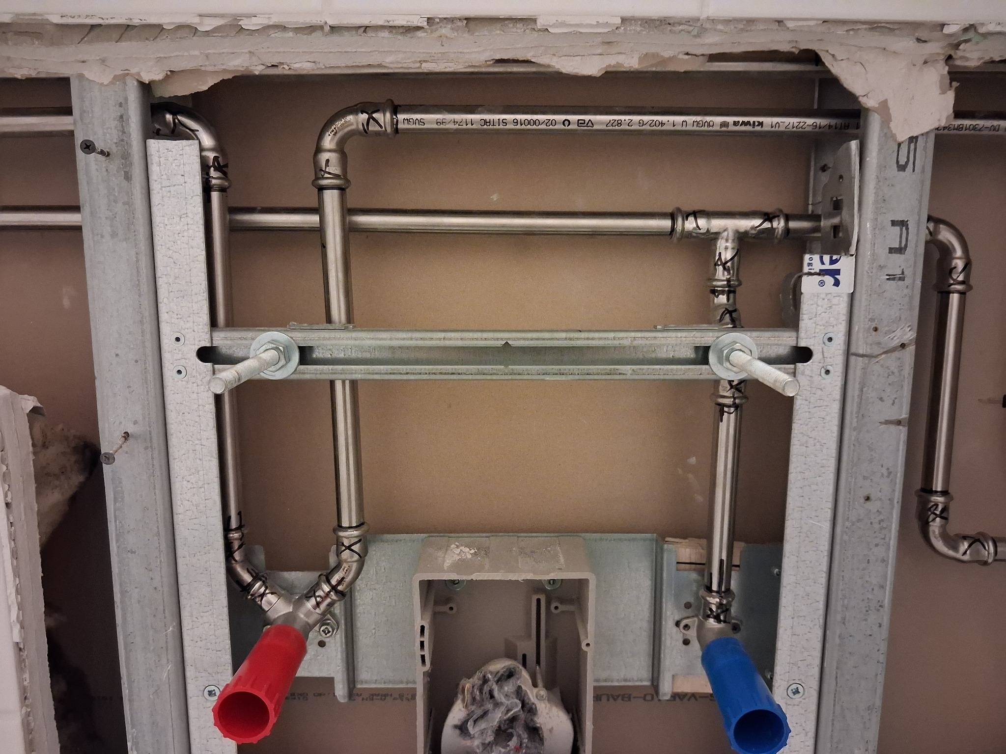 Szczegółowe ujęcie instalacji hydraulicznej w ścianie, z widocznymi rurami ze stali nierdzewnej, elementami mocującymi i przyłączami oznaczonymi kolorami czerwonym i niebieskim.