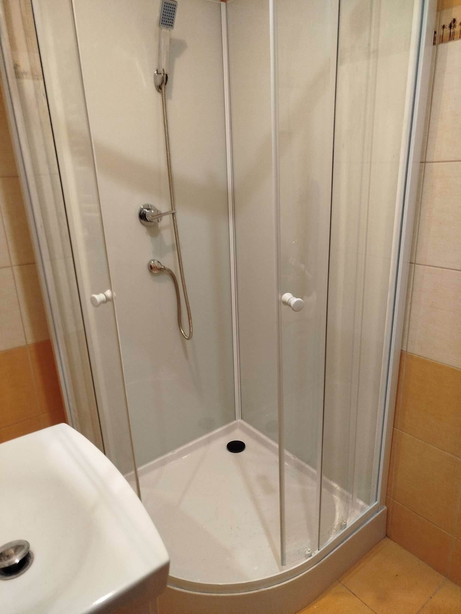 Kabina prysznicowa z brodzikiem i armaturą, widok z boku, częściowo widoczna umywalka. Beżowe płytki na ścianach. Zamontowana słuchawka prysznicowa.