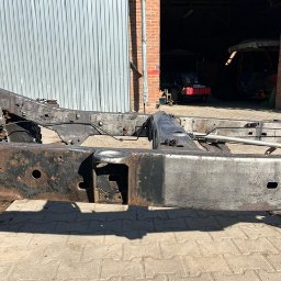 Jakub Puchała JMP - Studio - Rama pojazdu po czyszczeniu, widoczne ślady korozji, zarys garażu w tle. Elementy metalowe o nieregularnej powierzchni. Światło dzienne.