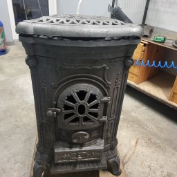 Jakub Puchała JMP - Studio - Żeliwny piec wolnostojący z dekoracyjnymi zdobieniami, stojący na podkładce w warsztacie. Widoczne detale drzwiczek i ażurowej góry pieca.