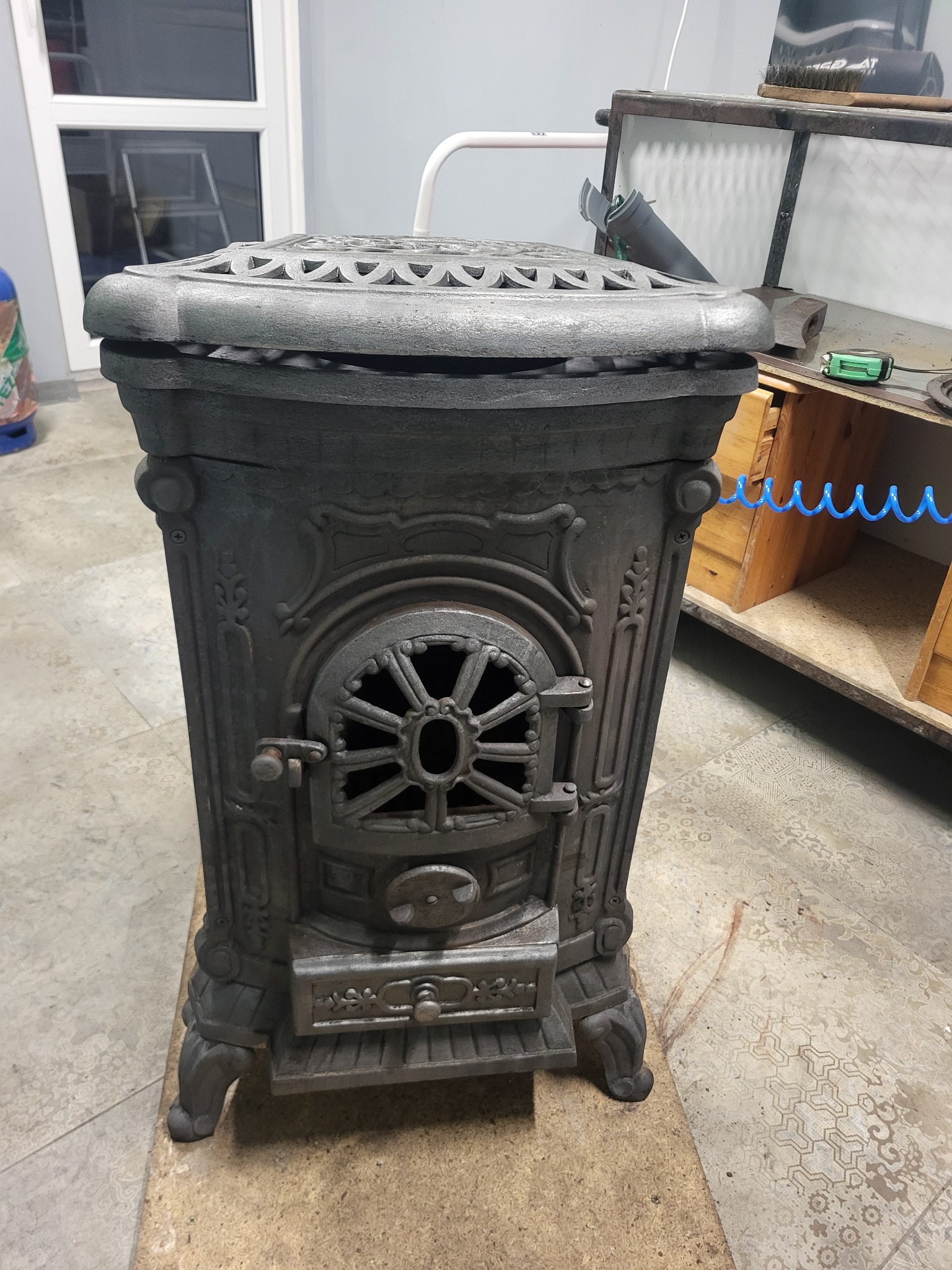 Żeliwny piec wolnostojący z dekoracyjnymi zdobieniami, stojący na podkładce w warsztacie. Widoczne detale drzwiczek i ażurowej góry pieca.