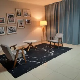 Pokój terapeutyczny z dwoma fotelami i stolikiem na czarnym dywanie z białym wzorem marmuru, oświetlony lampą stojącą, ściany zdobią zdjęcia w ramkach.