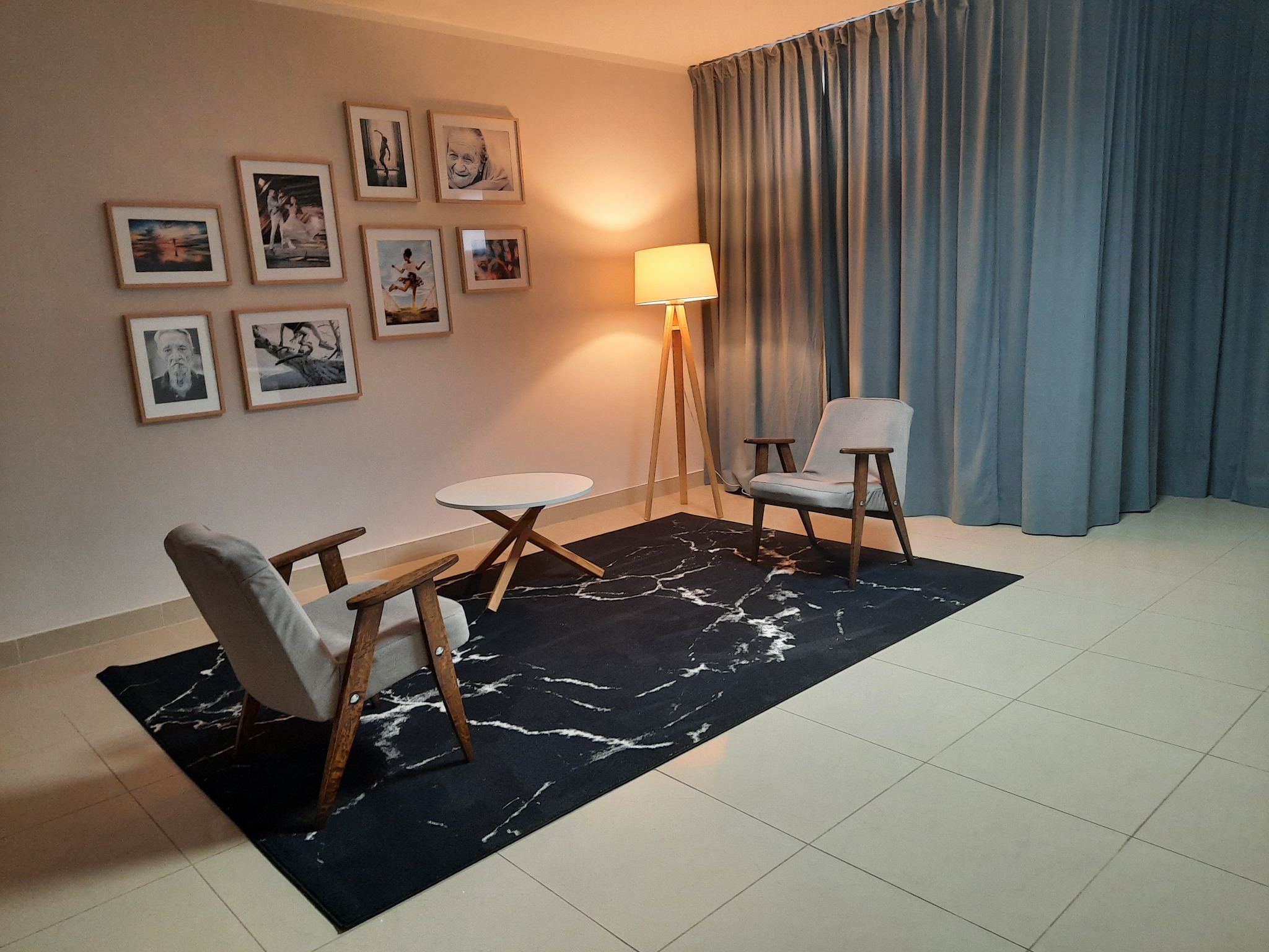 Pokój terapeutyczny z dwoma fotelami i stolikiem na czarnym dywanie z białym wzorem marmuru, oświetlony lampą stojącą, ściany zdobią zdjęcia w ramkach.