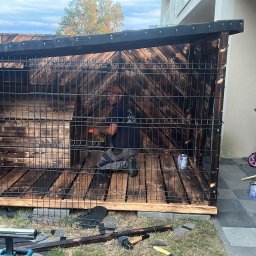 PKUBI-BAU Piotr Kubiciel - Mężczyzna wewnątrz drewnianej konstrukcji z siatką, malujący drewno na ciemny kolor, widoczne narzędzia i puszka farby, na zewnątrz domu.