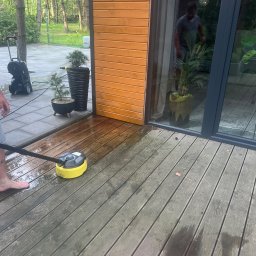 PKUBI-BAU Piotr Kubiciel - Czyszczenie drewnianego tarasu myjką ciśnieniową z okrągłą szczotką, widoczny fragment stopy osoby obsługującej, w tle dom z drewnianą elewacją i dużym oknem.