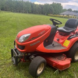 PKUBI-BAU Piotr Kubiciel - Czerwony traktorek ogrodowy Kubota GR2120 na trawiastym terenie, z widocznym lasem w tle i fragmentem domu.