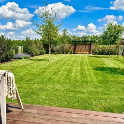 PKUBI-BAU Piotr Kubiciel - Perfekcyjnie utrzymany trawnik z widocznymi pasami po koszeniu, otoczony zielenią, z drewnianym tarasem i huśtawkami hamakowymi w tle, całość skąpana w słonecznym świetle pod błękitnym niebem...