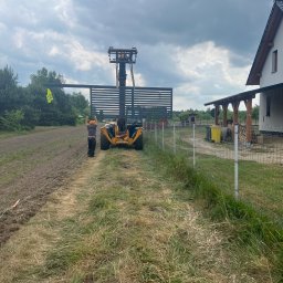 PKUBI-BAU Piotr Kubiciel - Montaż panelu ogrodzeniowego z pomocą podnośnika teleskopowego, pracownik w kamizelce odblaskowej i pomarańczowej czapce, widok na częściowo ukończone ogrodzenie panelowe i dom w tle.