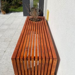 PKUBI-BAU Piotr Kubiciel - Drewniana ławka z wbudowaną donicą, wykonana z pionowych listew w ciepłym odcieniu brązu, umieszczona na szarym bruku w pobliżu białej ściany budynku.