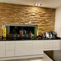 PKUBI-BAU Piotr Kubiciel - Nowoczesna kuchnia z dekoracyjną ścianą z drewnianych paneli, czarnym blatem, białymi szafkami, szklanymi pojemnikami z cytrynami i jabłkami oraz motywem brzozy na ścianie.