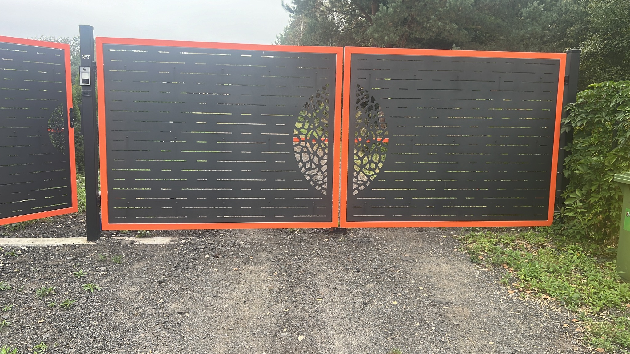 Nowoczesna, czarna brama panelowa z pomarańczową ramą i ażurowym wzorem liścia, zamontowana na posesji z widocznym otoczeniem zieleni.