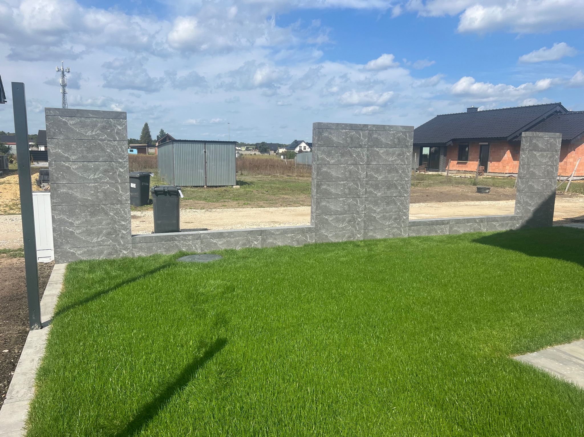 Fragment ogrodzenia panelowego z szarego betonu imitującego kamień, widok na trawnik, w tle budynek w trakcie budowy i blaszak.