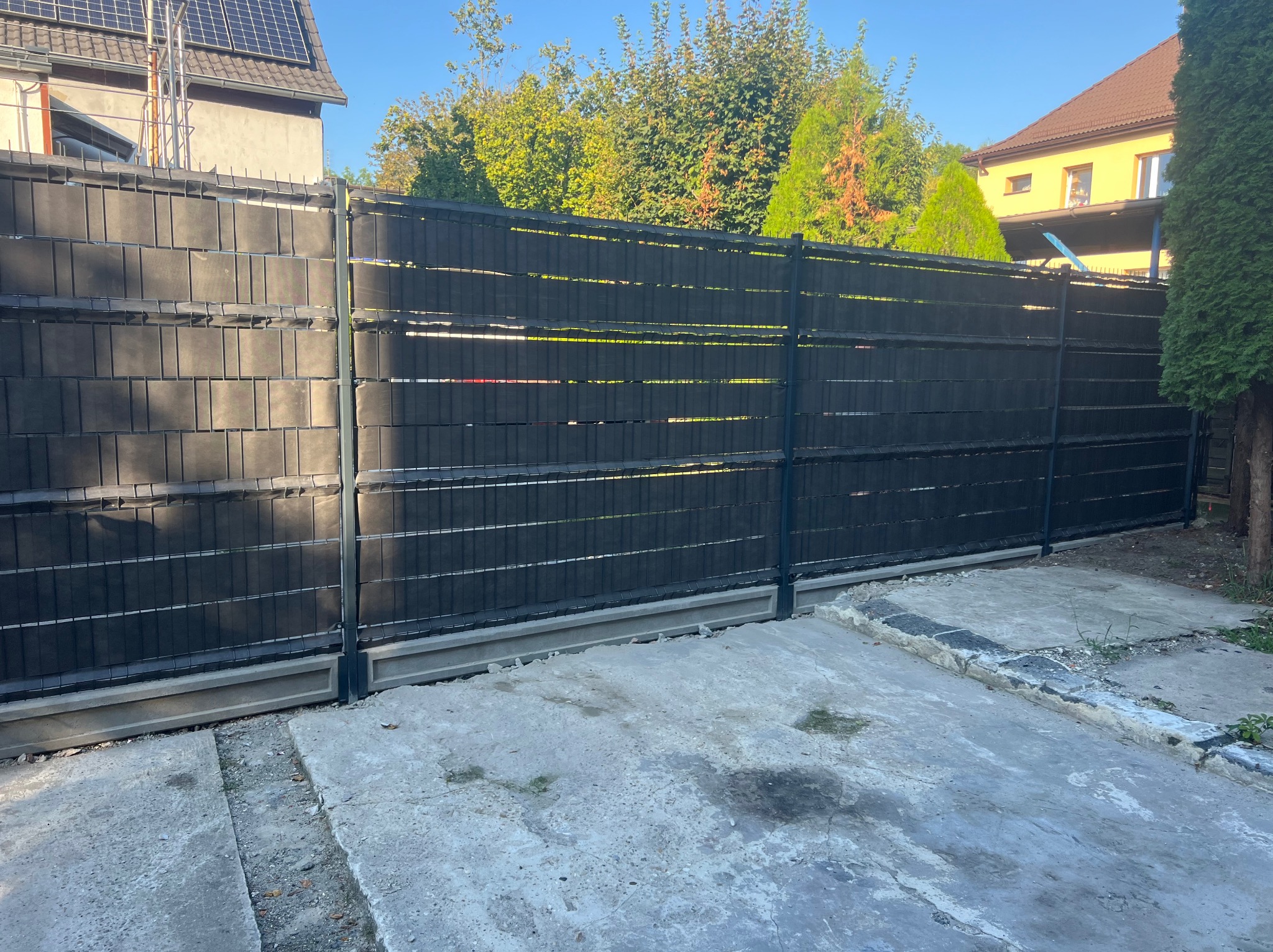 Nowoczesne, czarne ogrodzenie panelowe z betonową podmurówką, widoczne na tle zieleni drzew i budynku z panelami słonecznymi na dachu.