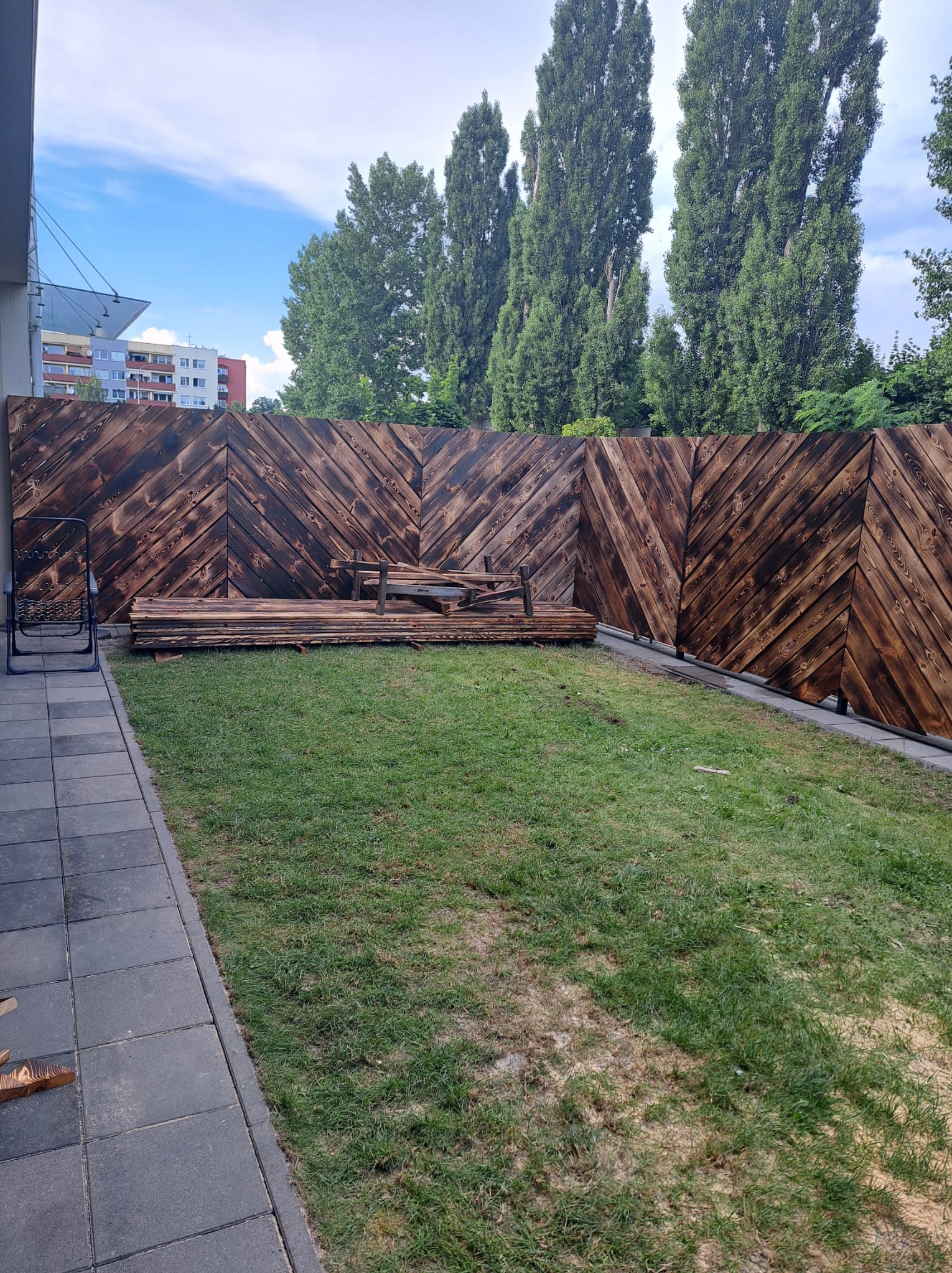 Nowoczesne ogrodzenie z ciemnego drewna ułożonego w jodełkę, z przygotowanymi deskami obok i krzesłem na tle trawnika, w tle wysokie drzewa i budynek.