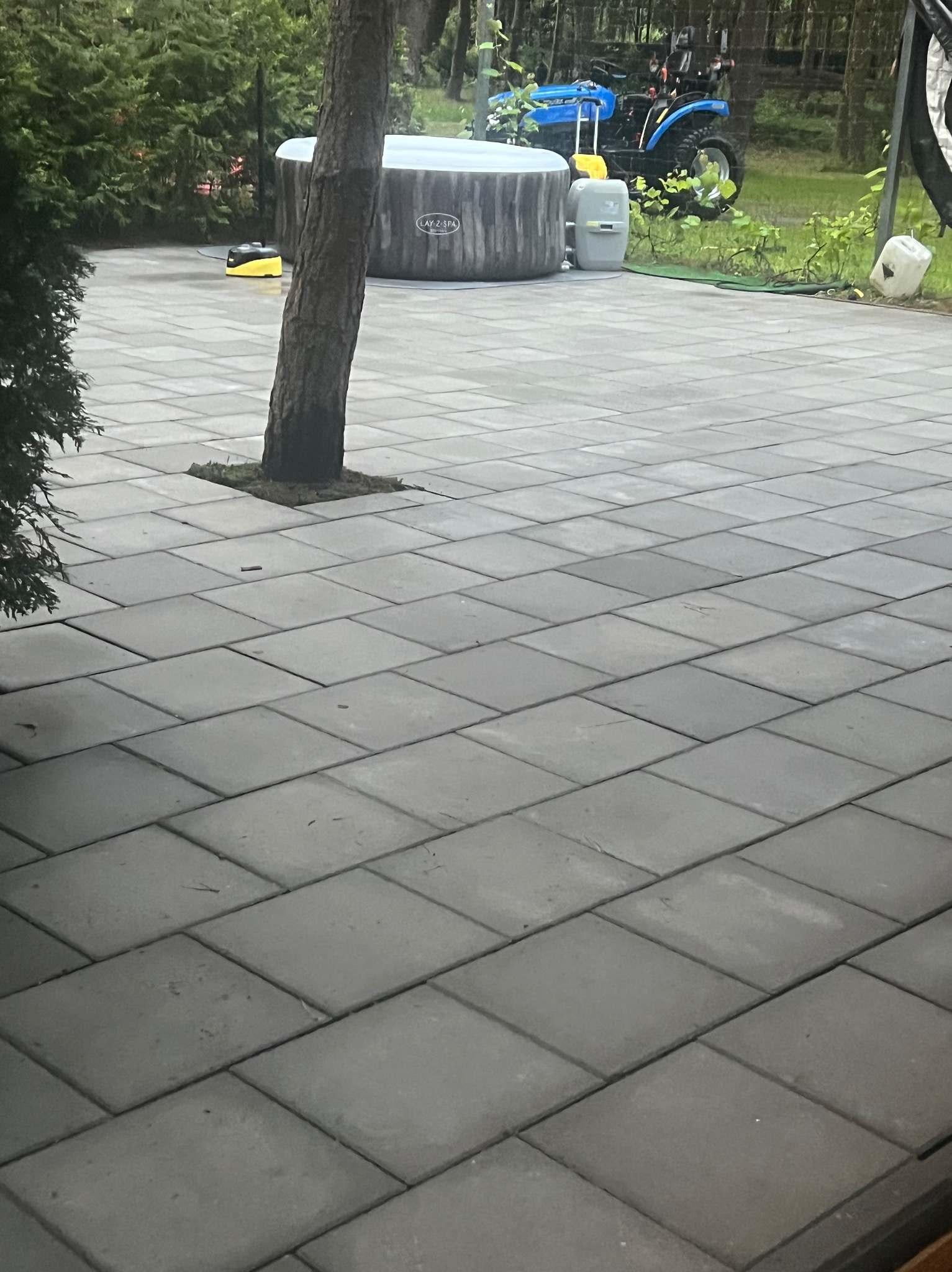 Szara kostka brukowa na posesji z rosnącym drzewem, w tle widoczny basen Lay-z-Spa i niebieski traktor ogrodowy.