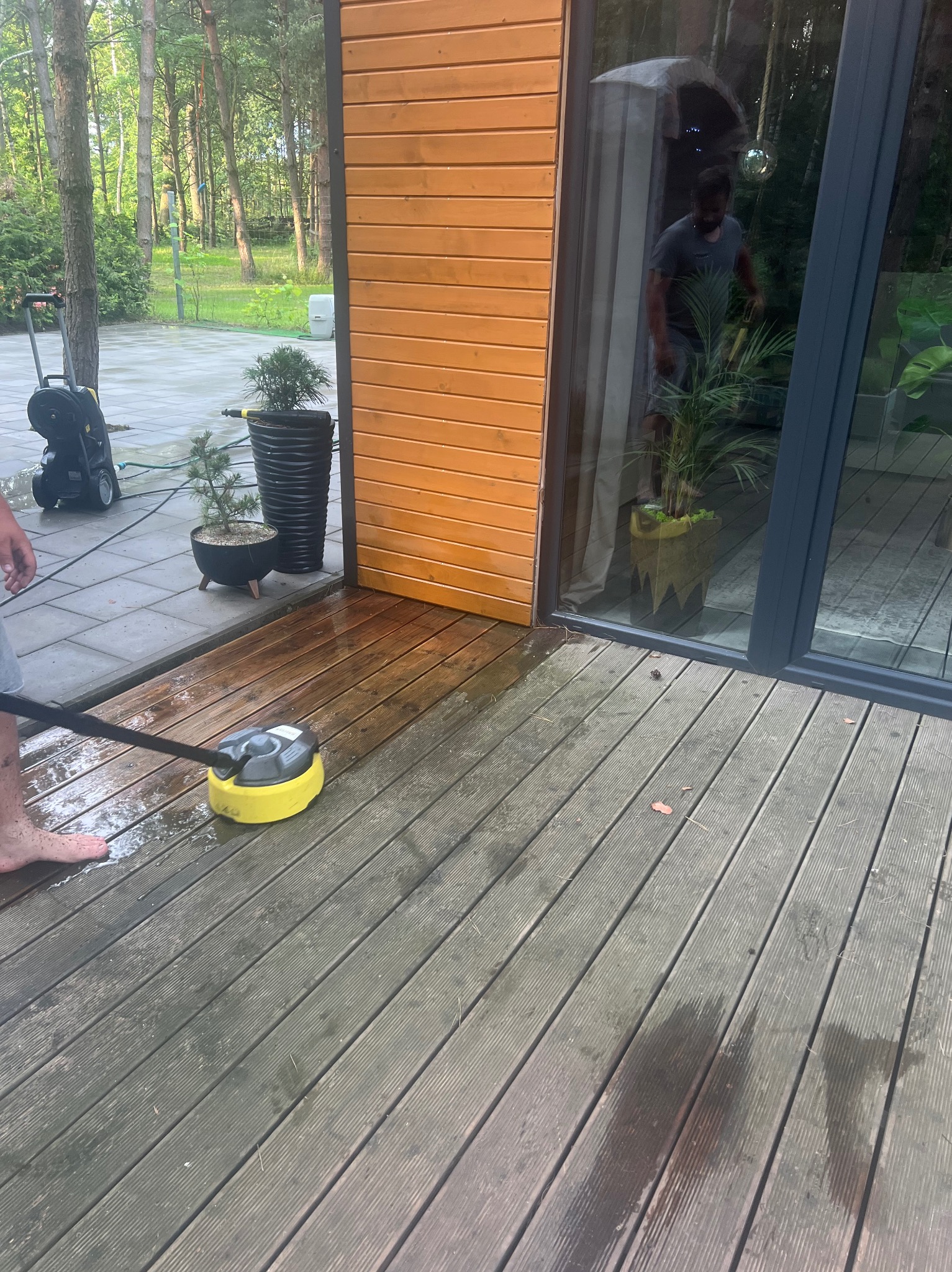 Czyszczenie drewnianego tarasu myjką ciśnieniową z okrągłą szczotką, widoczny fragment stopy osoby obsługującej, w tle dom z drewnianą elewacją i dużym oknem.