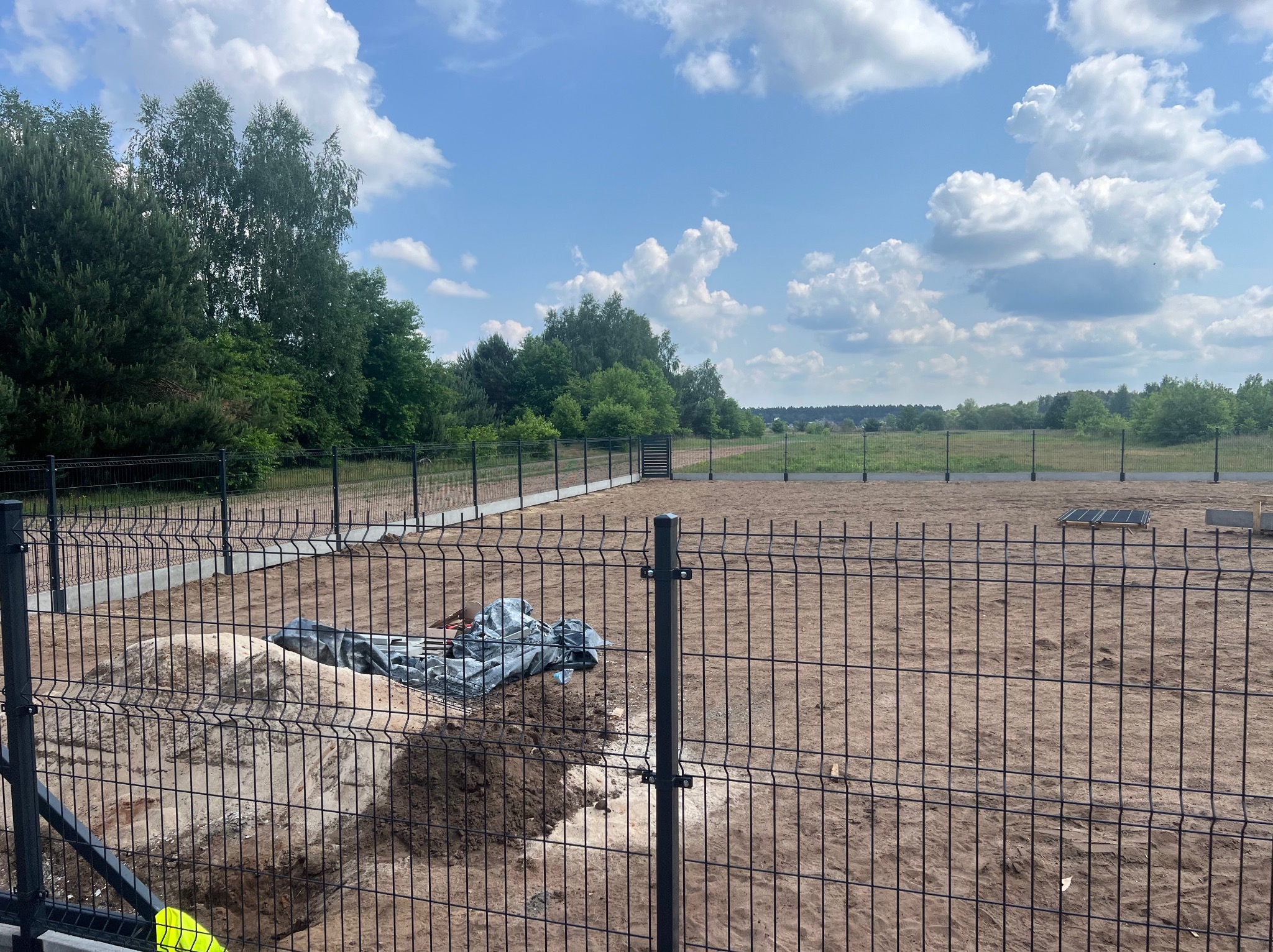 Nowo postawione, czarne ogrodzenie panelowe na piaszczystym terenie z widocznymi wykopami i stertą piachu przykrytą folią, w tle łąka i las pod błękitnym niebem z chmurami.