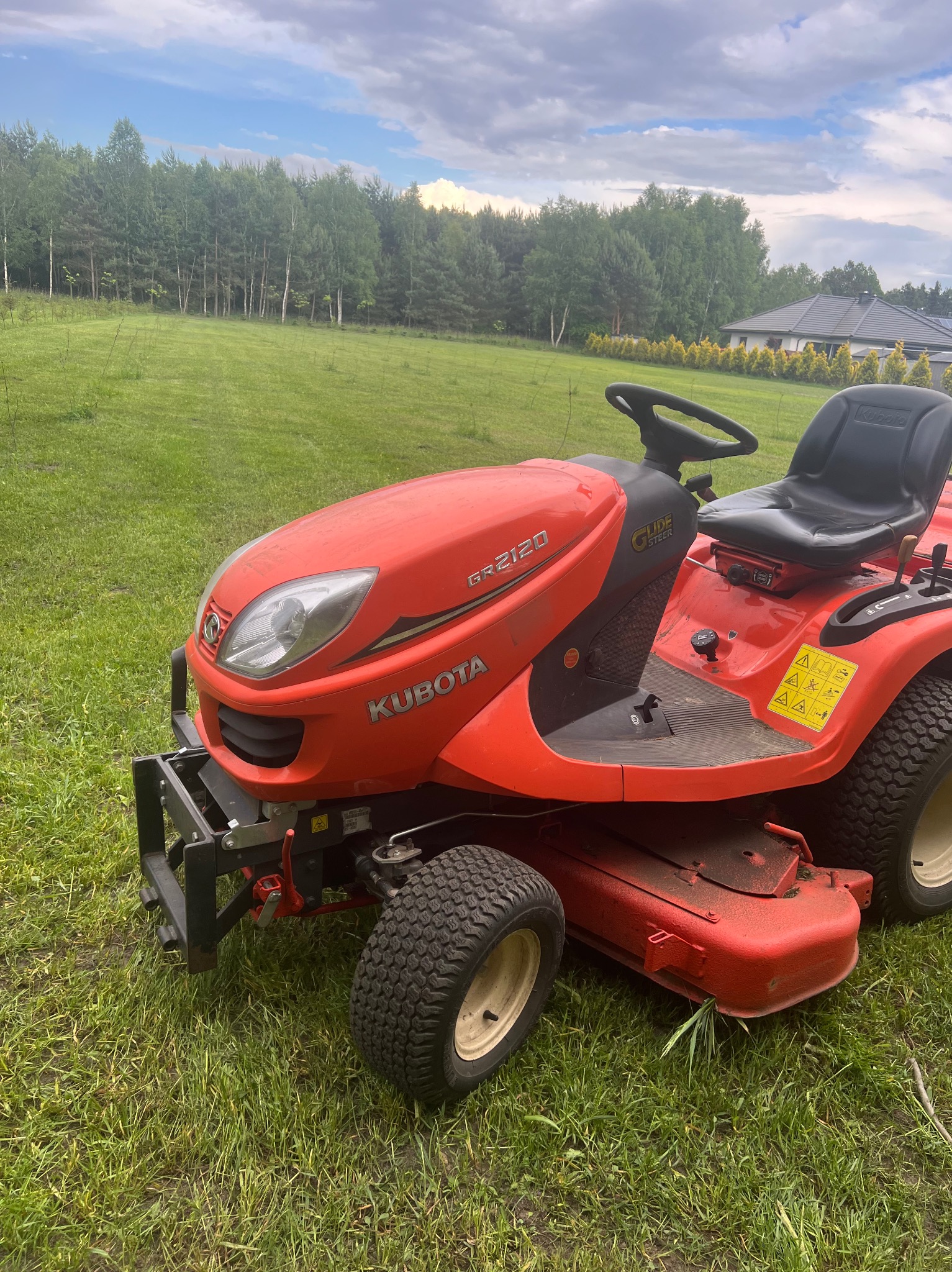 Czerwony traktorek ogrodowy Kubota GR2120 na trawiastym terenie, z widocznym lasem w tle i fragmentem domu.