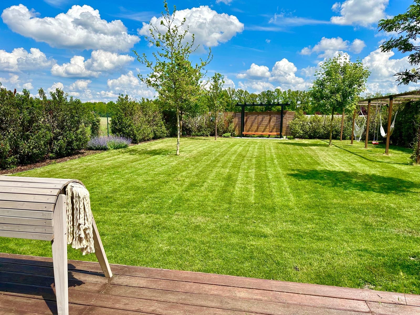 Perfekcyjnie utrzymany trawnik z widocznymi pasami po koszeniu, otoczony zielenią, z drewnianym tarasem i huśtawkami hamakowymi w tle, całość skąpana w słonecznym świetle pod błękitnym niebem...