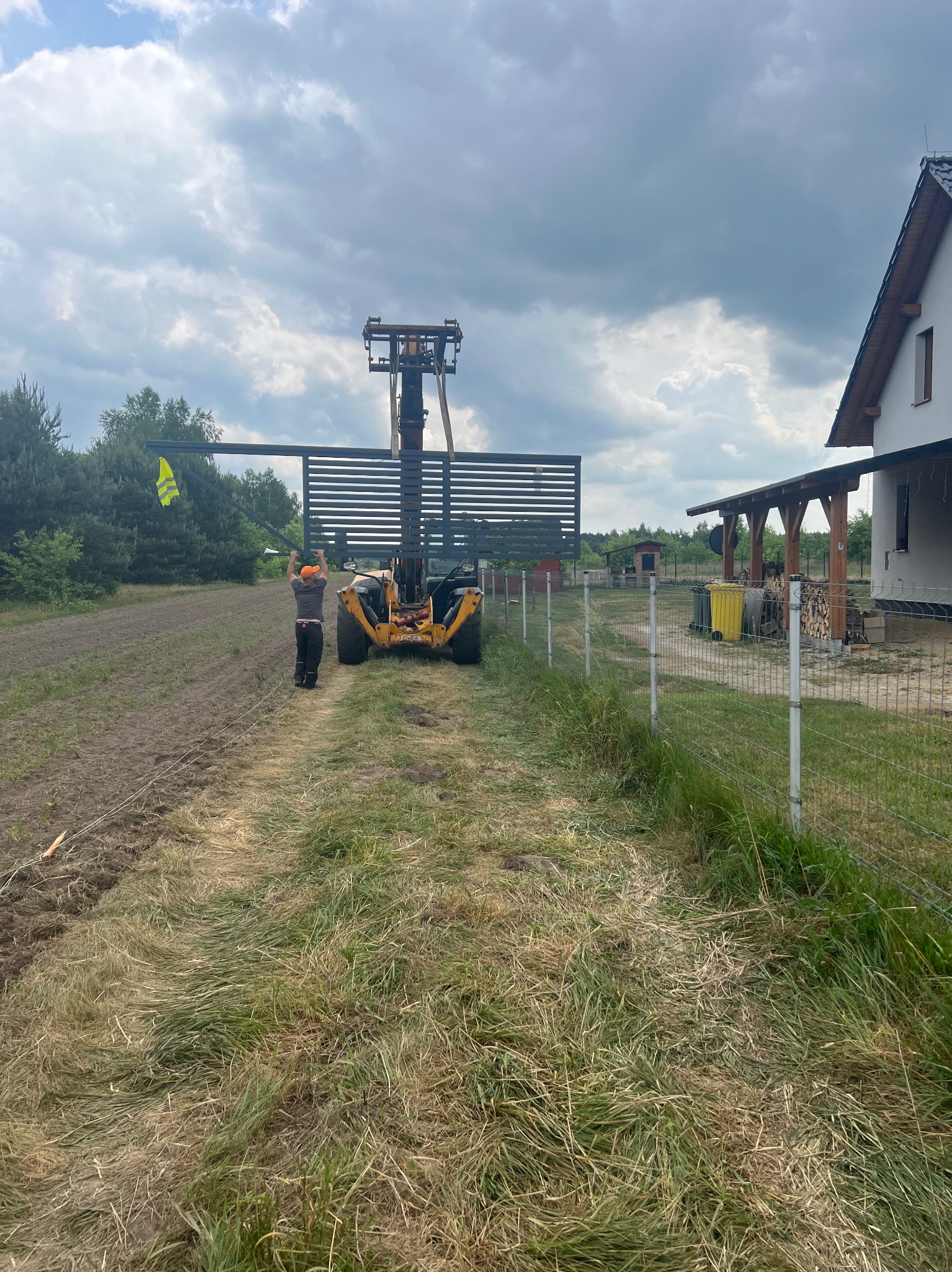 Montaż panelu ogrodzeniowego z pomocą podnośnika teleskopowego, pracownik w kamizelce odblaskowej i pomarańczowej czapce, widok na częściowo ukończone ogrodzenie panelowe i dom w tle.