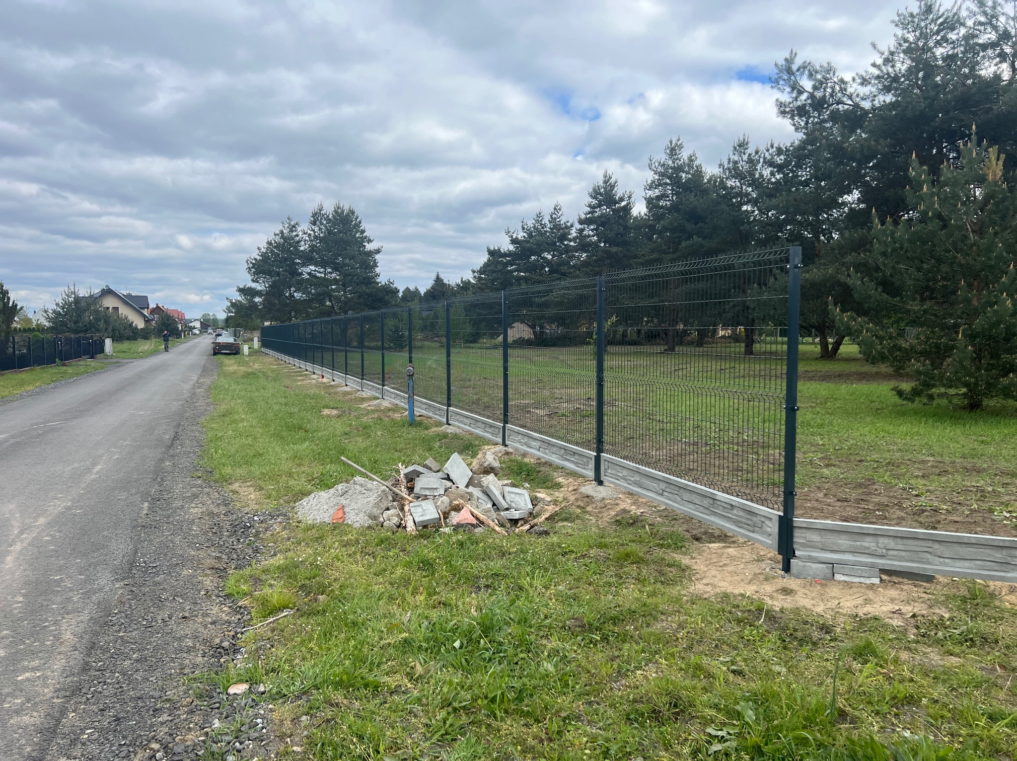 Nowo postawione ogrodzenie panelowe z podmurówką betonową, widoczne pozostałości po montażu, droga i zieleń w tle.
