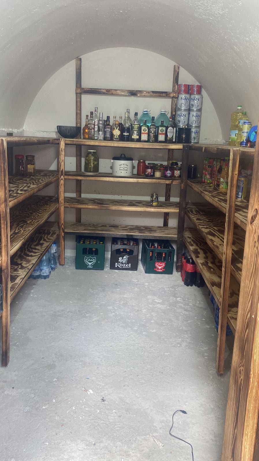 Domowa spiżarnia z drewnianymi regałami, wypełniona słoikami, butelkami, napojami i żywnością, umieszczona w pomieszczeniu o sklepionym suficie.